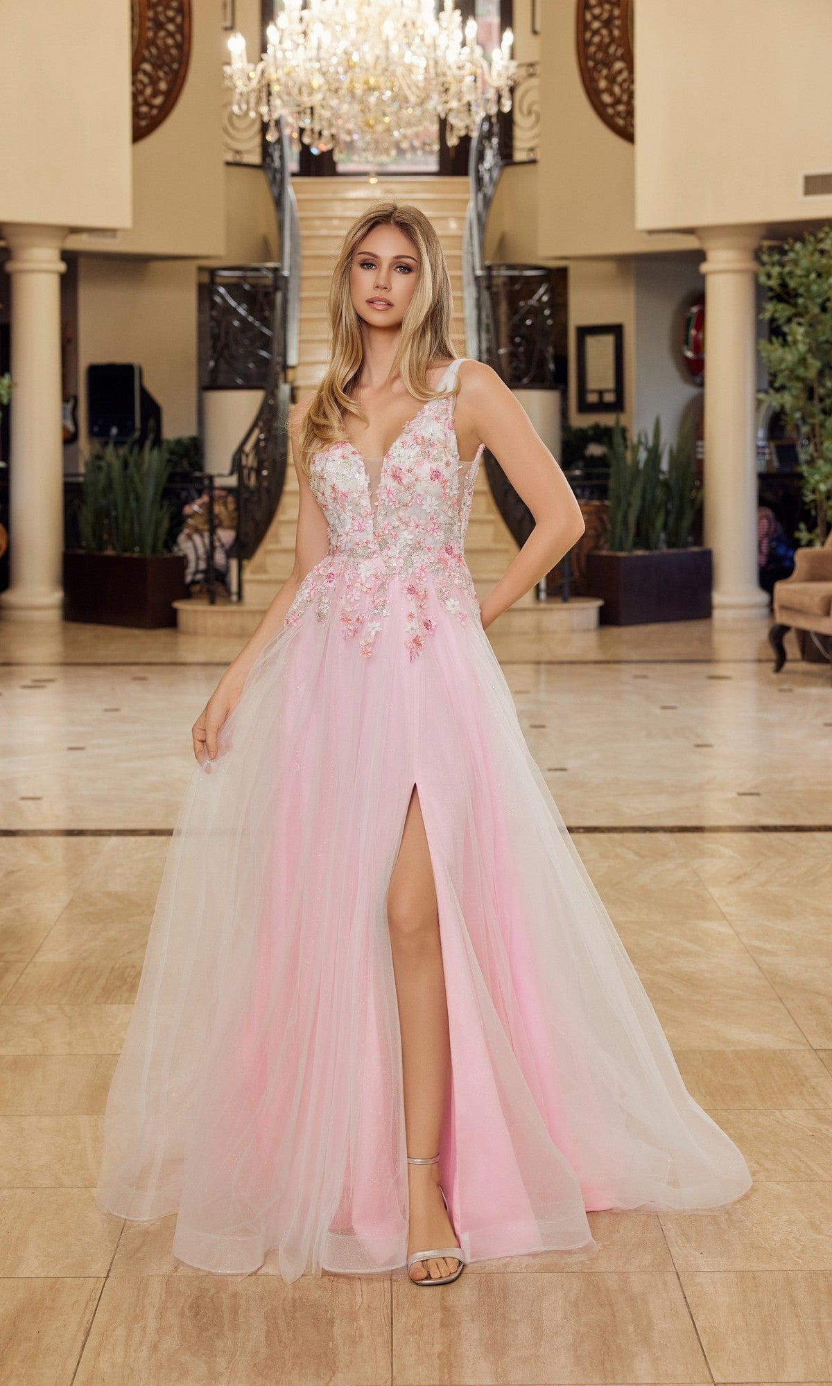 A woman wears the Nox Anabel Y1799 long prom dress in baby pink with floral appliques and a front slit, standing indoors before a staircase and chandelier.